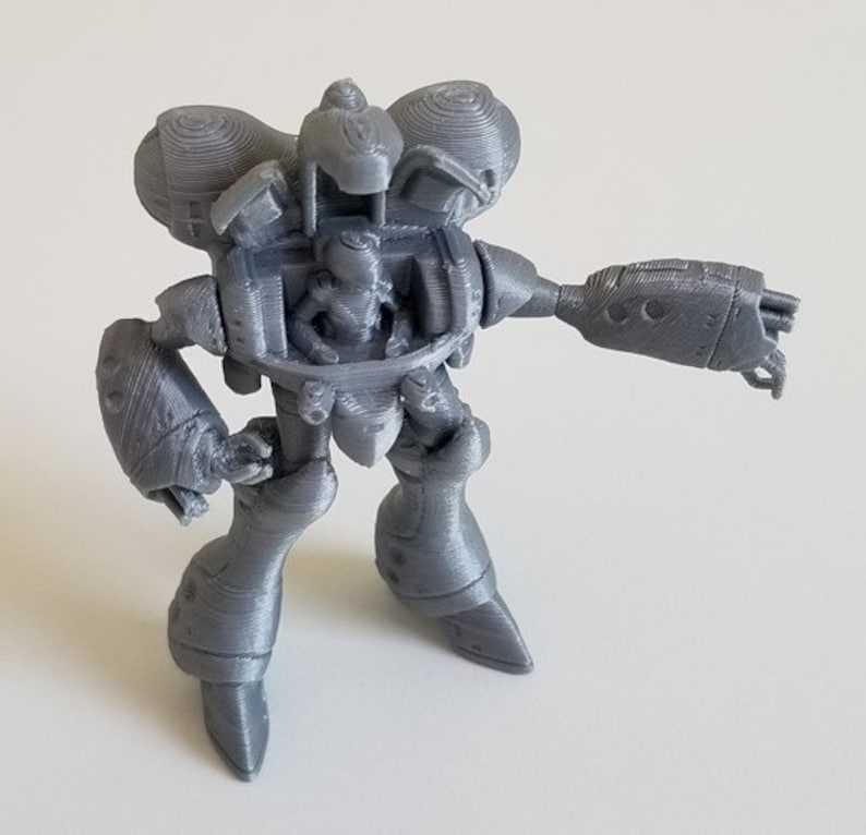 May include: A gray 3D printed robot toy with a detailed design. The robot has a cockpit, two arms, and two legs.