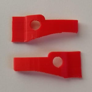 May include: Two bright red plastic parts, each with a rectangular base, a circular hole, and a tapered extension. The parts are likely components of a larger item or mechanism. The color is a vibrant, solid red.