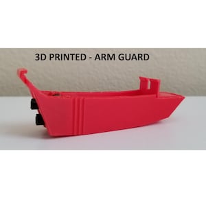 May include: A 3D-printed red arm guard. The arm guard is boat-shaped with a raised front and two black cylinders on the side. The text "3D PRINTED - ARM GUARD" is above the arm guard.