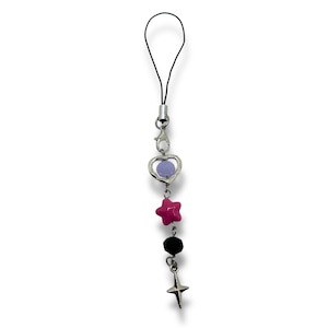 Puede incluir: Un charm decorativo para teléfono con un lazo de cordón negro y herrajes plateados. El charm presenta un colgante en forma de corazón con una cuenta morada, una estrella rosa, una cuenta negra y un charm de estrella plateada.