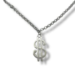 May include: Silver-toned necklace featuring a dollar sign pendant. The pendant is a stylized representation of the dollar symbol, suspended from a delicate chain. The necklace is displayed against a white background.