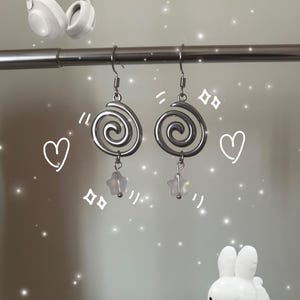 Pode incluir: Brincos espirais prateados com pingentes em forma de estrela. Os brincos estão pendurados numa barra de metal, com um coelho de pelúcia branco e fones de ouvido brancos no fundo. Design moderno e único.