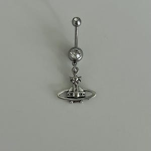 May include: Silver-toned belly button ring with a dangling charm. The charm features a small orb design with a ring around it. The top and middle balls are clear, faceted gems. The ring is designed for body piercing.