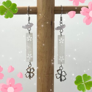 May include: A pair of dangle earrings featuring a silver-tone four-leaf clover charm. The earrings have a rectangular white accent with a floral design and a small pink accent at the top. The earrings are suspended from a silver hook.