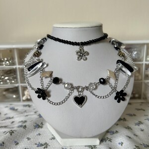 May include: A layered necklace with a silver chain, black beads, and clear crystals. The necklace features a black heart charm, two black flower charms, and a silver flower charm.