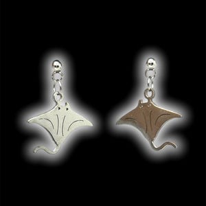 Puede incluir: Un par de pendientes colgantes plateados con un diseño de manta raya. Cada pendiente tiene un pequeño perno redondo conectado a una cadena que suspende la raya. Las rayas tienen una forma de ala detallada y una cola curva, una en plata y otra en tono bronce.