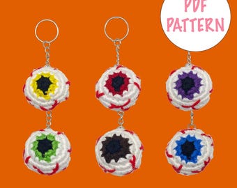 May include: Five crocheted eyeball keychains with different colored irises. The keychains are white with red veins and black pupils. The irises are yellow, red, purple, green, and blue. The keychains are hanging from silver key rings. The image includes the text "PDF PATTERN".