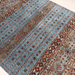 May include: A rectangular area rug featuring a light blue base with vertical bands of intricate geometric and floral patterns in shades of red, orange, brown, and cream. The design consists of repeating motifs arranged in stripes.