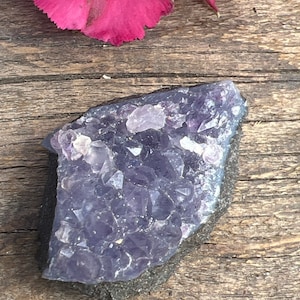 May include: A cluster of amethyst crystals in shades of purple and lavender, set in a dark gray matrix. The crystals are arranged in a natural, irregular shape, with some points and facets visible.