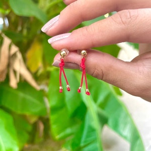 Puede incluir: Pendientes de oro con cordón rojo y detalles de cuentas doradas. Los pendientes tienen un diseño colgante con un detalle de nudo. El fondo es una vista borrosa de follaje verde.