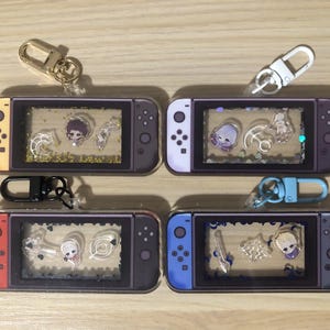 May include: Four rectangular acrylic shaker keychains shaped like handheld gaming consoles. Each features a clear window containing small floating charms and glitter. The controllers have different coloured side grips including yellow, white, red, and blue.