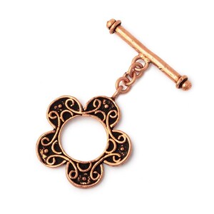 May include: A copper-toned toggle clasp featuring a flower-shaped ring with black recessed detailing and intricate scrollwork patterns. The clasp includes a matching straight bar component connected to the flower ring by two small circular metal links.