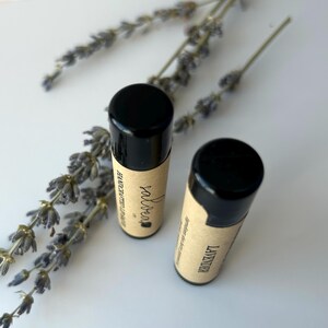 May include: Two black and brown lip balm tubes with labels that say "salvcrea" and "lavender" on a white surface with lavender flowers.