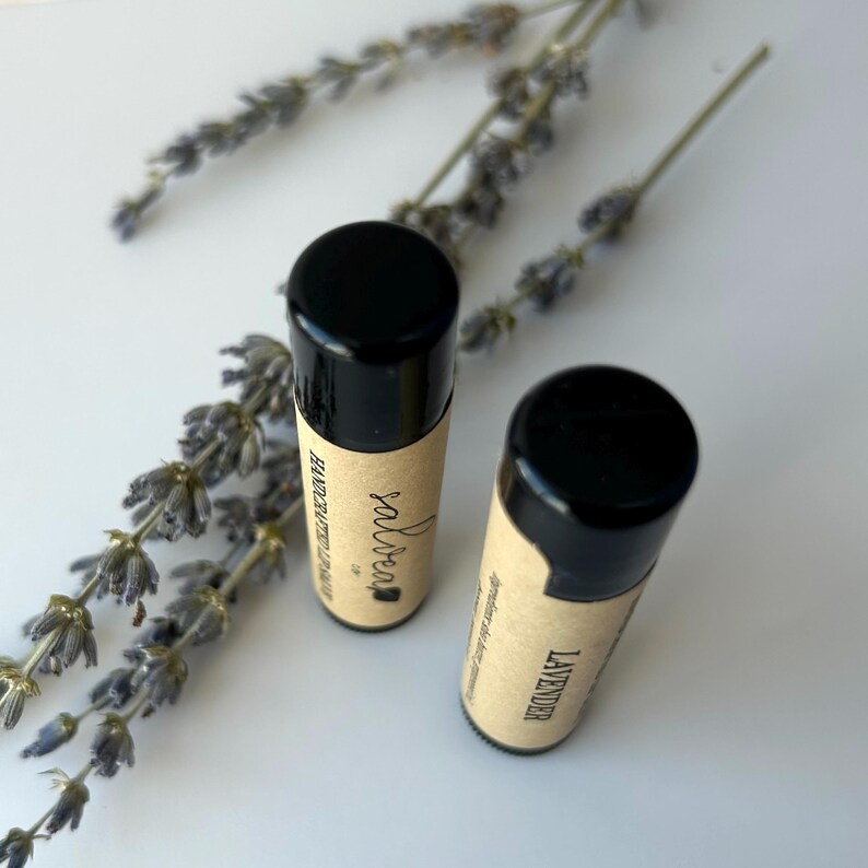 May include: Two black and brown lip balm tubes with labels that say "salvcrea" and "lavender" on a white surface with lavender flowers.