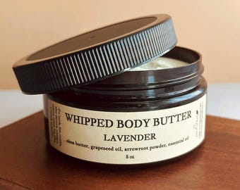 May include: A dark brown plastic jar of whipped body butter with a black ribbed lid resting at an angle. The cream-coloured label reads WHIPPED BODY BUTTER LAVENDER, listing shea butter, grapeseed oil, arrowroot powder, and essential oil, 8 oz.