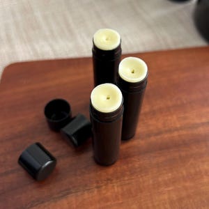 May include: Three black lip balm tubes with a white, waxy substance inside. The tubes are on a wooden surface.