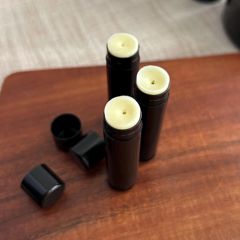 May include: Three black lip balm tubes with a white, waxy substance inside. The tubes are on a wooden surface.