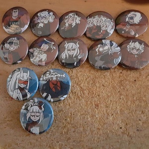 Puede incluir: Una colección de 12 pines redondas con varios personajes de dibujos animados. Los pines son coloridos y tienen un acabado brillante. Los personajes se representan en varias poses y expresiones. Los pines están dispuestos en un círculo sobre una superficie marrón.