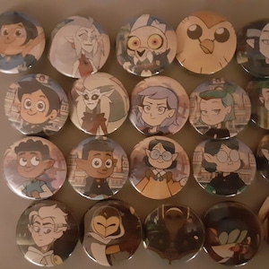 May include: A collection of 18 round buttons featuring characters from the animated series The Owl House. The buttons are colourful and feature various characters, including a young witch with purple hair, a young boy with brown hair, and an owl-like creature.
