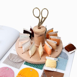 Op de afbeelding: Een houten draadorganizer met een centrale spoelhouder en een cirkelvormige basis met verschillende spoelen draad in tinten bruin, oranje en crème. Een gouden schaar is in de spoelhouder gestoken. De organizer ligt op een open boek.