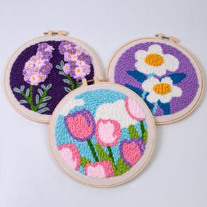 May include: Three floral embroidery hoops. One features purple flowers on a dark purple background, another has white flowers with yellow centers on a purple background, and the third has pink and purple tulips on a blue background.