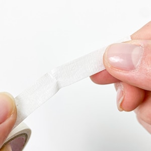 May include: A roll of white masking tape with a textured paper surface. The tape is being pulled from the roll, showing its matte finish and slightly fibrous, opaque appearance. The material is thin, flexible, and designed for adhesive applications.