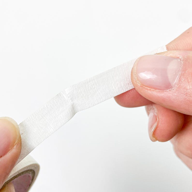 May include: A roll of white masking tape with a textured paper surface. The tape is being pulled from the roll, showing its matte finish and slightly fibrous, opaque appearance. The material is thin, flexible, and designed for adhesive applications.