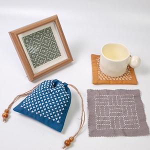 May include: A wooden framed textile art piece with a green geometric pattern, a blue drawstring pouch with white cross-stitch embroidery, a cream ceramic mug on an orange coaster, and a gray square coaster with white linear embroidery.