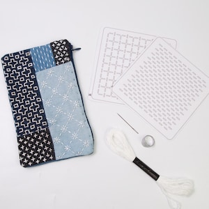 May include: A rectangular zippered pouch featuring blue and white geometric embroidery, accompanied by two white pattern templates, a single sewing needle, a metal thimble, and a skein of white embroidery thread for stitching projects.