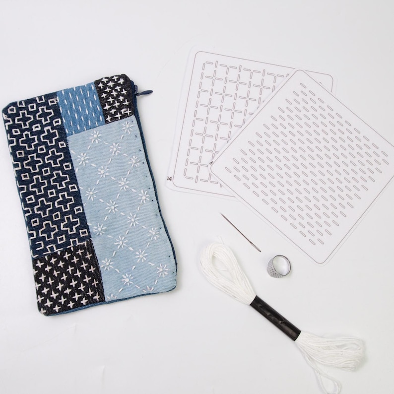 May include: A rectangular zippered pouch featuring blue and white geometric embroidery, accompanied by two white pattern templates, a single sewing needle, a metal thimble, and a skein of white embroidery thread for stitching projects.