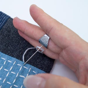 May include: A silver-colored open-sided thimble with a dimpled surface worn on a finger, positioned to push a long, thin sewing needle through a piece of dark blue denim fabric featuring white stitched grid patterns.