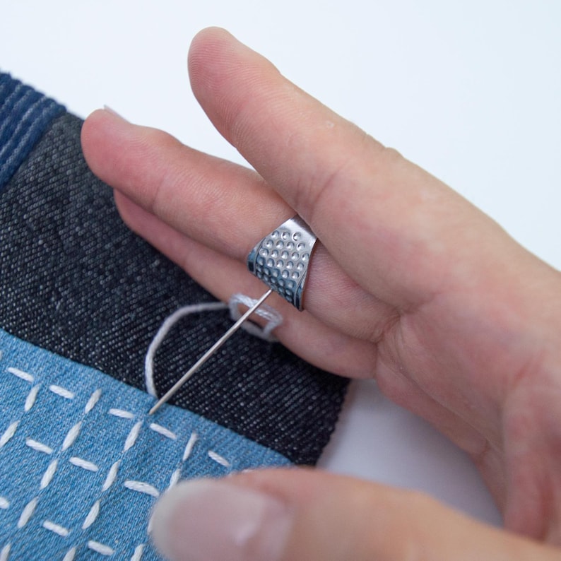 May include: A silver-colored open-sided thimble with a dimpled surface worn on a finger, positioned to push a long, thin sewing needle through a piece of dark blue denim fabric featuring white stitched grid patterns.