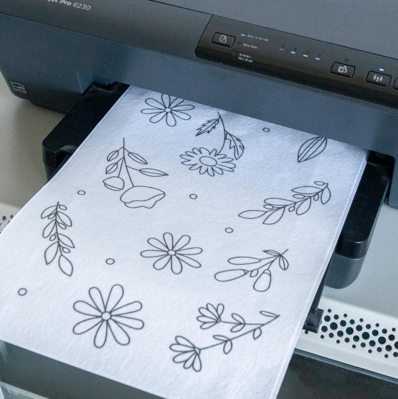 May include: A black inkjet printer with a sheet of white paper emerging from the tray. The paper features a black line art pattern of various flowers, leaves, and small circles scattered across the surface, creating a simple botanical design.