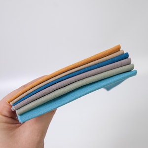 May include: A stack of five folded fabric swatches in a spectrum of colors including mustard yellow, tan, dark blue, light purple, and sage green, each featuring a visible woven texture and clean, straight edges stacked neatly on top of each other.