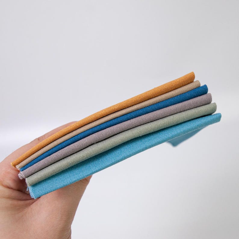 May include: A stack of five folded fabric swatches in a spectrum of colors including mustard yellow, tan, dark blue, light purple, and sage green, each featuring a visible woven texture and clean, straight edges stacked neatly on top of each other.