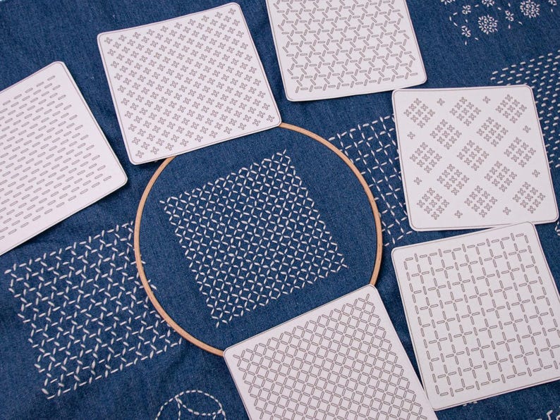 May include: A circular wooden embroidery hoop containing blue fabric with white stitched geometric patterns, surrounded by six square white templates featuring various black line-art grid designs for embroidery and needlework projects.