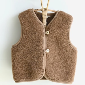 Può includere: Un gilet di pile marrone con chiusura a bottone. Il gilet è appeso a un appendiabiti di legno.