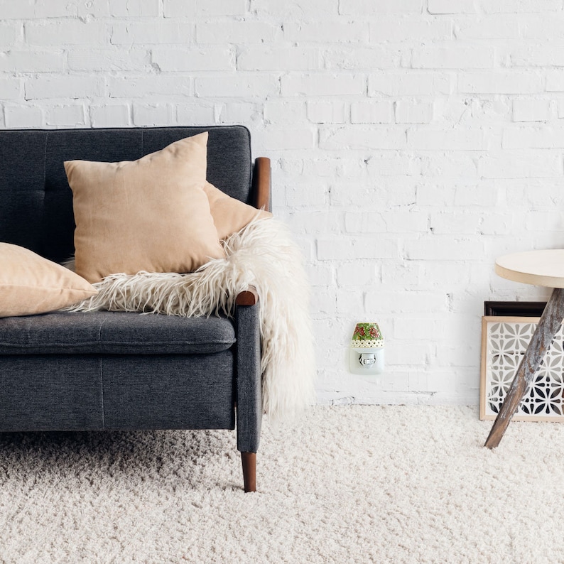 K&ouml;nnte beinhalten: Ein graues Sofa mit einer flauschigen wei&szlig;en Decke und beigen Kissen. Eine wei&szlig;e Nachtlampe mit floralem Design ist an einer Steckdose an einer wei&szlig;en Backsteinwand angeschlossen.