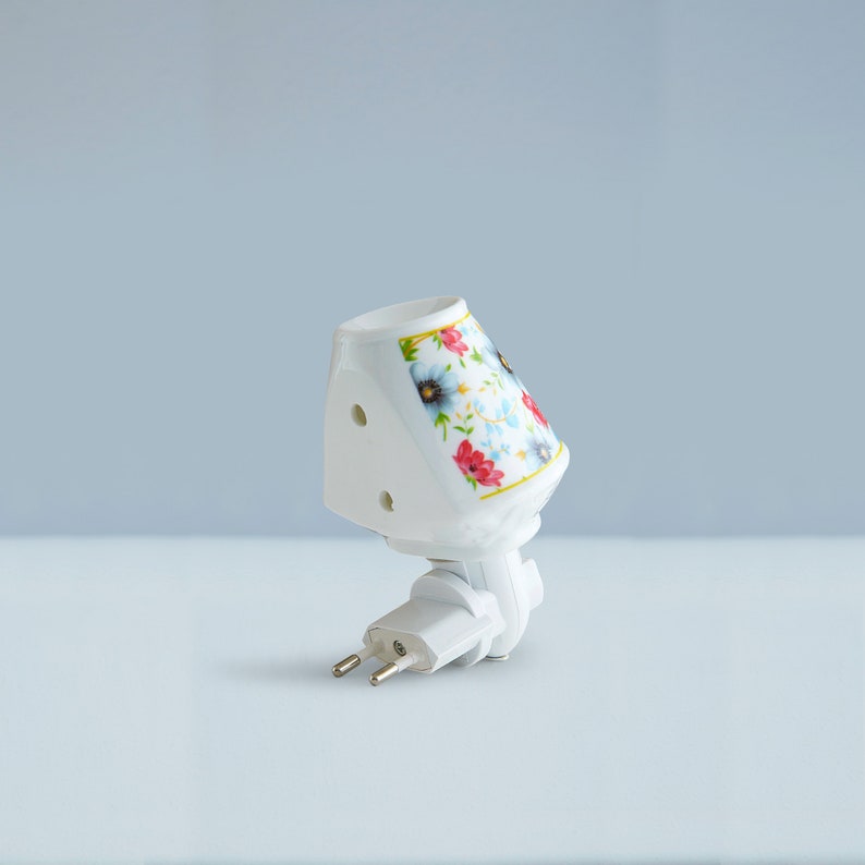 Pu&ograve; includere: Un piccolo faretto da notte bianco in ceramica con un design floreale. Il faretto ha una forma triangolare e una spina a due poli.