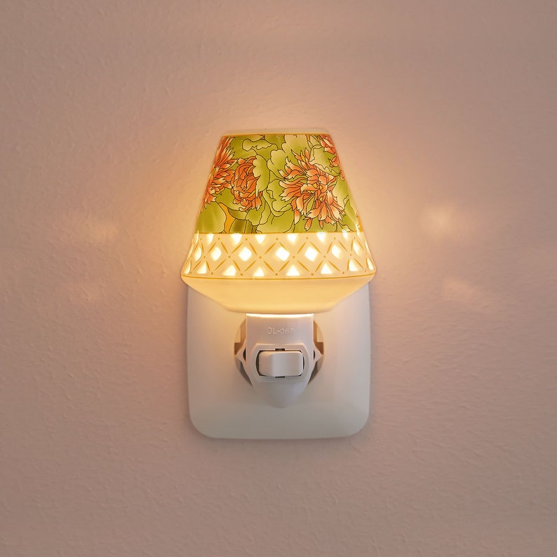 K&ouml;nnte beinhalten: Ein wei&szlig;er keramischer Nachtlicht-Stecker mit einem floralen Design auf dem Schirm. Der Schirm hat ein gr&uuml;nes und orangefarbenes Blumenmuster mit einem wei&szlig;en Gitterdesign. Das Nachtlicht ist in eine normale Steckdose gesteckt.