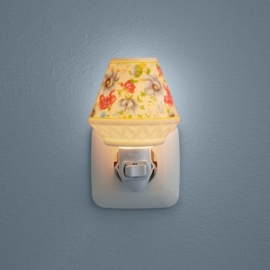 Könnte beinhalten: Ein weißes Keramik-Nachtlicht mit einem Blumendesign auf dem Schirm. Das Nachtlicht ist an eine Steckdose angeschlossen und hat einen Schalter an der Seite.