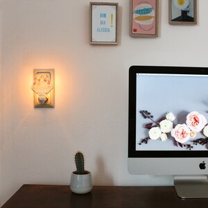 Pu&ograve; includere: Una luce notturna bianca con un designo floreale collegata a una presa a muro bianca. La luce notturna &egrave; accesa e proietta un bagliore caldo. Una piccola pianta di cactus &egrave; in un vaso bianco su un tavolo di legno davanti a un monitor di computer. Il monitor del computer mostra uno sfondo desktop con fiori bianchi e bacche.