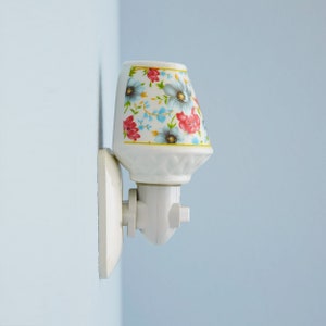 Pu&ograve; includere: Un lampadario da parete in ceramica bianca con un motivo floreale. Il lampadario ha un cavo bianco e una spina bianca.