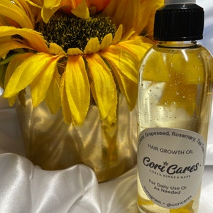 May include: A clear glass bottle of hair growth oil with a black cap, next to a gold vase with yellow sunflowers. The label reads "Hair Growth Oil Cori Cares" with instructions for use. The background is white.