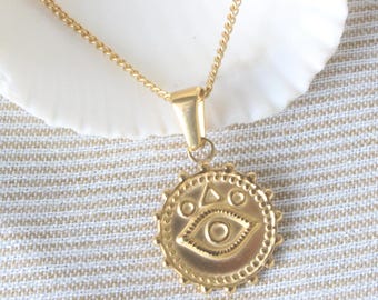 May include: Gold necklace with a round pendant featuring a detailed evil eye design. The pendant has a circle of small beads around the edge and a triangle inside the eye.