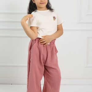 May include: A young person wearing a white t-shirt with a floral embroidery design and pink wide-leg trousers.
