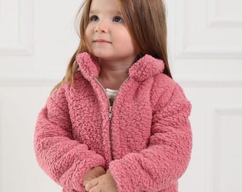 May include: A pink fuzzy jacket with a zipper closure and a pocket on the front. The jacket is made of a soft, plush material.