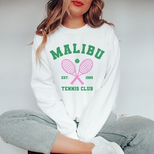 K&ouml;nnte beinhalten: Wei&szlig;er Pullover mit einem gr&uuml;nen und rosafarbenen Grafikdesign, auf dem "Malibu Tennis Club Est. 1985" mit zwei rosafarbenen Tennisschl&auml;gern und einem gr&uuml;nen Tennisball steht.