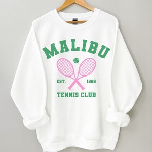K&ouml;nnte beinhalten: Wei&szlig;er Pullover mit einem gr&uuml;nen und rosafarbenen Grafikdesign, auf dem "MALIBU TENNIS CLUB EST. 1985" mit zwei rosafarbenen Tennisschl&auml;gern und einem gr&uuml;nen Tennisball steht.