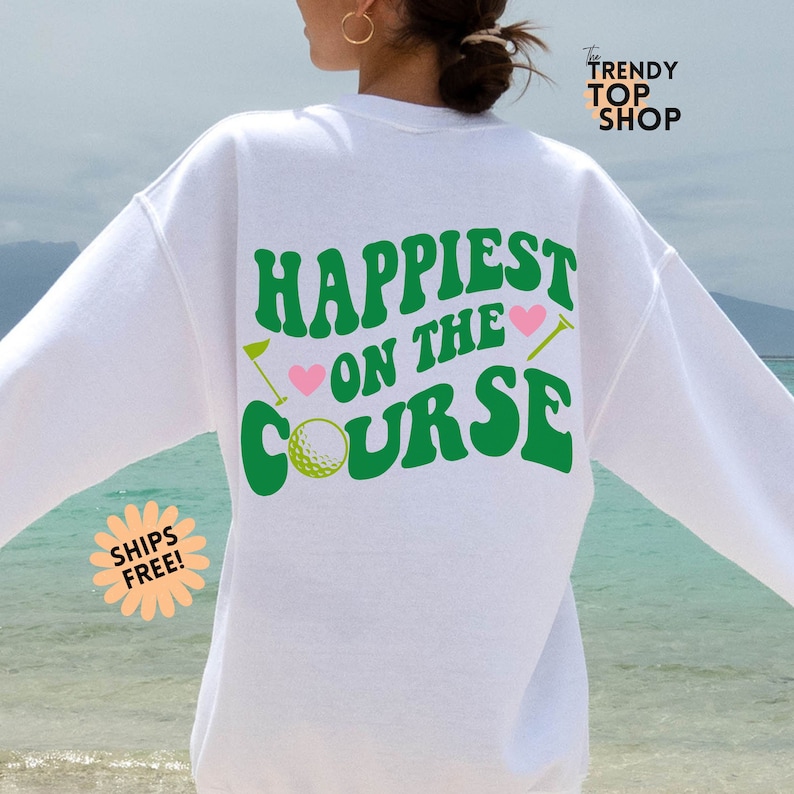 May include: White sweatshirt with green text that says "Happiest on the Course" with a golf ball and golf club graphic.